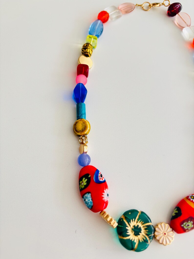 May include: A colorful beaded necklace with a variety of shapes and sizes. The necklace features red, blue, green, and gold beads, with a gold clasp. The focal point includes red oval beads with floral designs.
