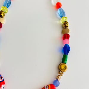 May include: A colorful beaded necklace with a gold clasp. The necklace features a variety of beads in red, blue, yellow, green, and pink. Some beads have floral patterns. The necklace is on a white background.