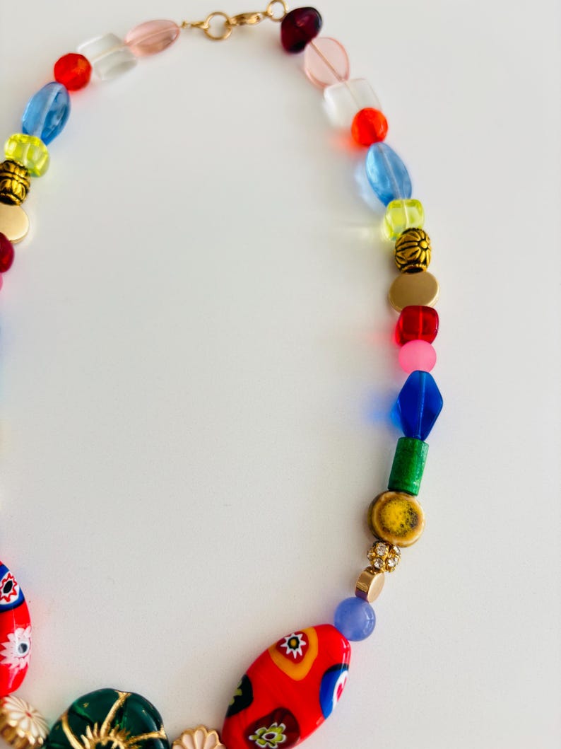 May include: A colorful beaded necklace with a gold clasp. The necklace features a variety of beads in red, blue, yellow, green, and pink. Some beads have floral patterns. The necklace is on a white background.