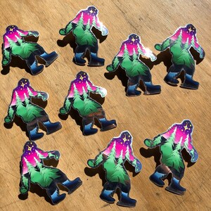 May include: Nine colorful stickers of a Bigfoot with a forest scene inside its head. The stickers are holographic and have a shiny finish. The Bigfoot is green, pink, and blue with a star in the sky.