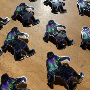 May include: A set of nine silver holographic stickers featuring a Bigfoot silhouette with a purple, green, and black aurora borealis design.