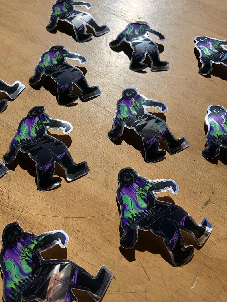 May include: A set of nine silver holographic stickers featuring a Bigfoot silhouette with a purple, green, and black aurora borealis design.
