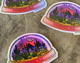 May include: Three colorful stickers with a desert scene inside a snow globe. The stickers feature a purple, pink, and blue sky with a UFO, cacti, and mountains. The text on the stickers reads "PHOENIX AZ" and "@misskittytattoo".