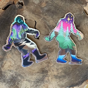May include: Two holographic stickers featuring a Bigfoot silhouette. One sticker has a purple, blue, and green design with a crescent moon and stars. The other sticker has a pink, green, and blue design with a star and pine trees.