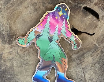May include: A colorful sticker of a Bigfoot with a starry sky and pine trees inside its silhouette. The sticker has a holographic finish and the text "@MISSSKITTYTATTOO" at the bottom.