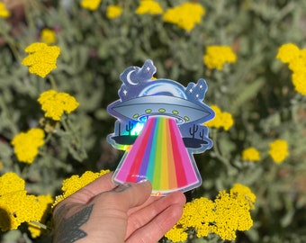 May include: A holographic sticker of a UFO shooting a rainbow beam of light. The sticker is in front of a background of yellow flowers.