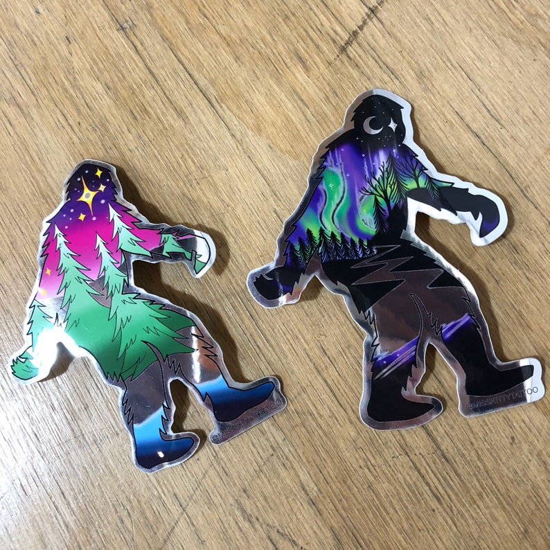May include: Two silver foil stickers of Bigfoot, one with a green, pink, and blue aurora borealis design and the other with a purple and blue aurora borealis design.