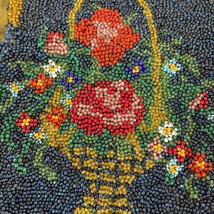 May include: A close-up of a beaded artwork depicting a basket overflowing with colorful flowers. The basket is made of gold beads and the flowers are made of red, pink, white, and green beads.