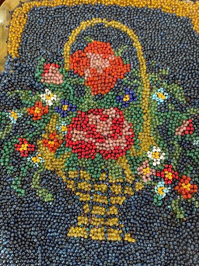 May include: A close-up of a beaded artwork depicting a basket overflowing with colorful flowers. The basket is made of gold beads and the flowers are made of red, pink, white, and green beads.