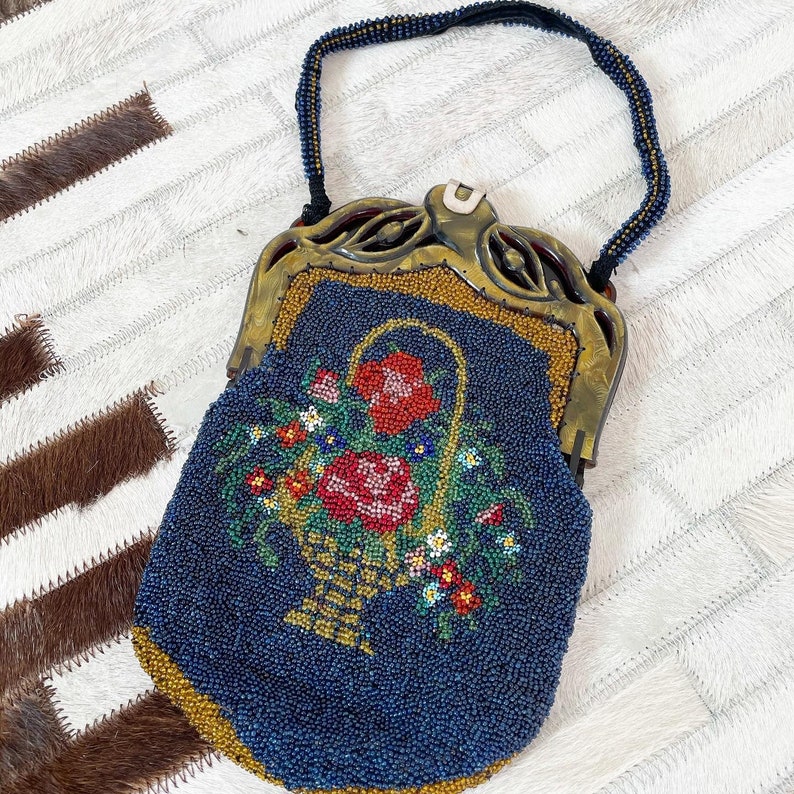 May include: A vintage beaded purse with a floral design. The purse is blue with gold accents and features a basket of flowers with red, pink, and yellow blooms.