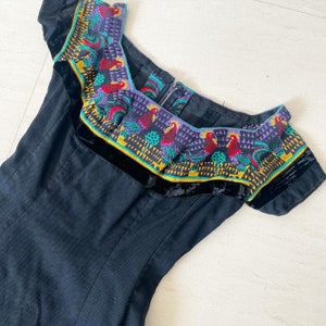 Pode incluir: Um vestido preto com um decote bordado de cores vibrantes que representa pássaros e padrões geométricos. O decote é guarnecido com veludo preto.