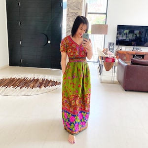 Puede incluir: Una mujer lleva un vestido maxi verde con estampado floral. El vestido tiene un corpiño ajustado y una falda completa. El vestido está acentuado con un cinturón negro y flores rosas.