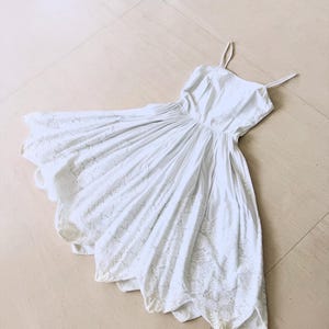 May include: A white sundress with spaghetti straps and a gathered waist. The dress features a scalloped hemline with embroidered floral detailing. The fabric appears to be lightweight, suitable for warm weather.