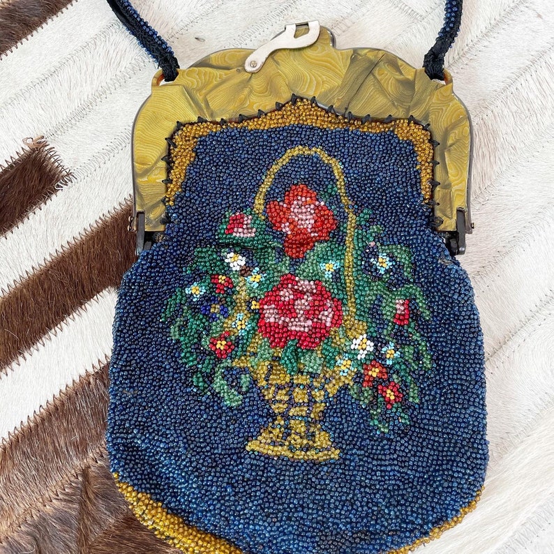 May include: A vintage beaded purse with a floral design. The purse is blue with gold accents and features a basket of flowers with red, pink, and white blooms.