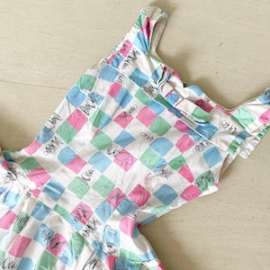 Puede incluir: Un vestido sin mangas con un patrón de cuadros en rosa, azul y verde. El vestido tiene un lazo en la parte posterior del cuello y un fondo blanco.