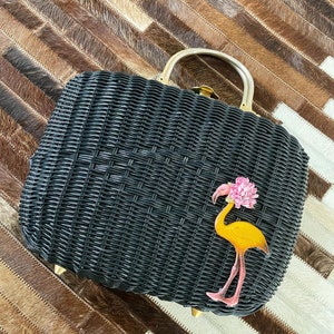 May include: Black woven wicker handbag with a gold handle and accents. The handbag features a yellow flamingo with a pink flower on its head. The bag is sitting on a brown and white patterned surface.
