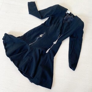 Puede incluir: Un vestido negro con mangas largas y un dobladillo con volantes. El vestido tiene una cremallera en la parte delantera y adornos plateados.