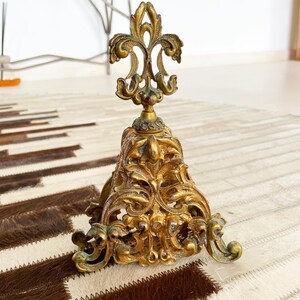 Puede incluir: Un tapón de botella de metal dorado con un diseño ornamentado e intrincado. El tapón presenta un remate de flor de lis y una base con un patrón decorativo en forma de rollo.