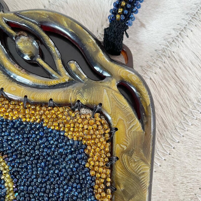 May include: A gold and black beaded purse with a unique eye-shaped design. The purse is made of a textured material and has a beaded strap.