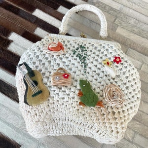 Puede incluir: Bolso de mano blanco de crochet con una variedad de adornos coloridos, incluyendo una rana, una guitarra, una flor y un pájaro. El bolso tiene un asa superior y un cierre de broche.