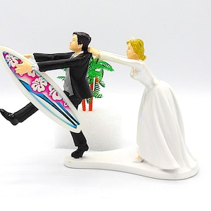 May include: Wedding cake topper featuring a groom in a black suit running with a surfboard decorated with pink and blue floral designs. A bride in a white gown tries to stop him. Palm trees are in the background.