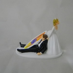 May include: A white wedding cake topper featuring a bride and groom. The groom is lying on his back with a surfboard under him. The bride is standing and holding the groom's hand. The surfboard has a colorful design.