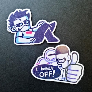 May include: Two stickers with cartoon illustrations. One shows a figure reclining with a stern expression. The other features a figure giving a thumbs up with the text "I beat OFF!" in a speech bubble. Both have a blue and white colour scheme.