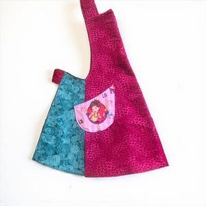 May include: A handmade tote bag with a unique design. The bag features a vibrant mix of patterns and colors, including a teal floral print, a magenta leopard print, and a pink pocket with a cartoon character. The bag has a single strap.