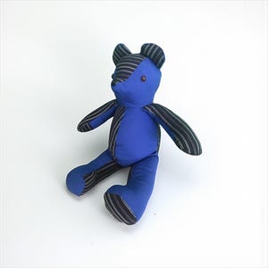 May include: A handmade blue teddy bear with striped fabric accents on the ears, arms, and legs. The bear has a round, blue body and a small, brown button eye. The bear is sitting on a white surface.