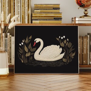 May include: A black and gold framed print of a white swan with a red beak, swimming in a pond with green and gold foliage. The print is on a shelf with books and a globe.