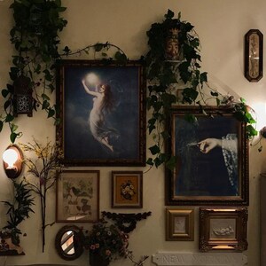 May include: A wall gallery featuring a variety of framed artwork, including a painting of a woman holding a glowing orb, a painting of a hand holding a feather, and a framed floral print. The wall is decorated with ivy and other greenery.