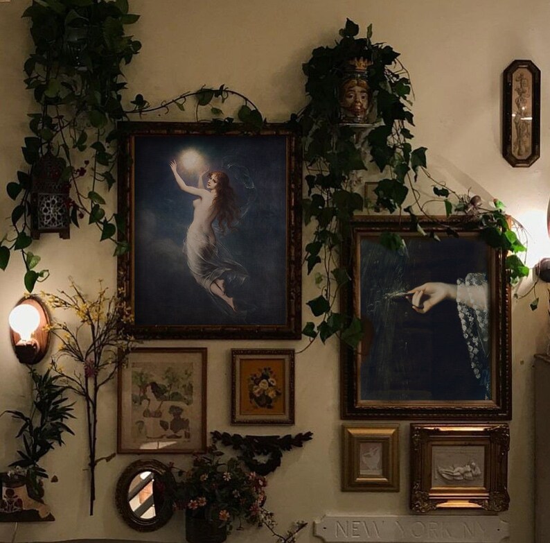 May include: A wall gallery featuring a variety of framed artwork, including a painting of a woman holding a glowing orb, a painting of a hand holding a feather, and a framed floral print. The wall is decorated with ivy and other greenery.