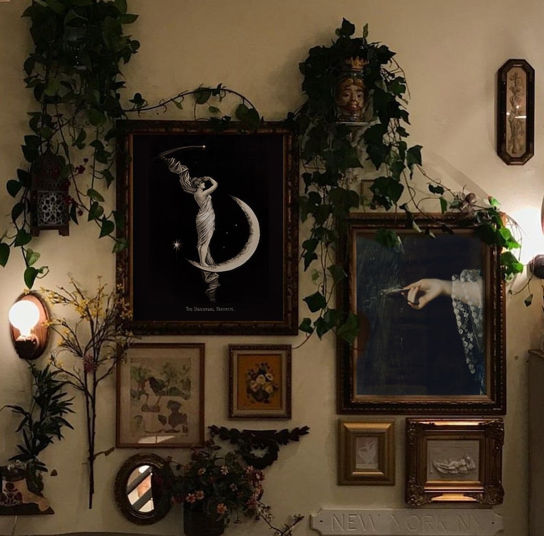 Puede incluir: Una galer&iacute;a de pared con una variedad de obras de arte enmarcadas, que incluyen una ilustraci&oacute;n en blanco y negro de una mujer de pie sobre una luna creciente, una pintura de una mano sosteniendo una pluma y una impresi&oacute;n bot&aacute;nica enmarcada. La galer&iacute;a est&aacute; decorada con vegetaci&oacute;n y un peque&ntilde;o espejo.