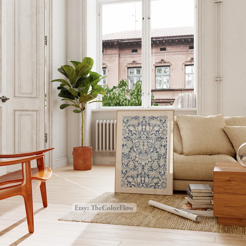May include: A blue and white floral print art print in a gold frame hanging on a white wall in a living room. The print is surrounded by a beige rug, a wooden chair, a beige sofa, and a wooden side table with books on it.