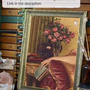 May include: A framed painting of a woman's hand resting on a book, with a vase of pink roses in the background. The frame is dark gold with ornate details. The painting is set on a table with a book and a vase of flowers.