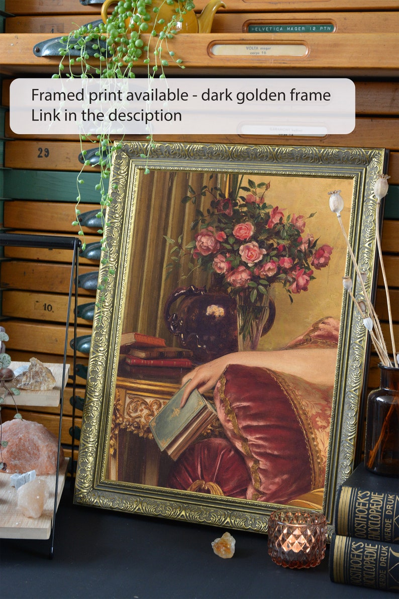 May include: A framed painting of a woman's hand resting on a book, with a vase of pink roses in the background. The frame is dark gold with ornate details. The painting is set on a table with a book and a vase of flowers.