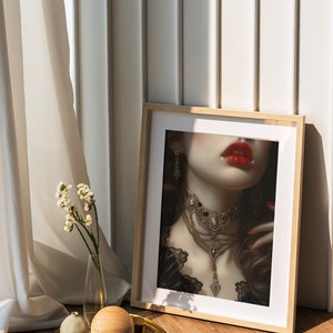 Puede incluir: Una impresi&oacute;n enmarcada de la cara de una mujer con l&aacute;piz labial rojo y un collar de oro grande. La impresi&oacute;n est&aacute; en un marco de madera y se apoya contra una pared blanca.