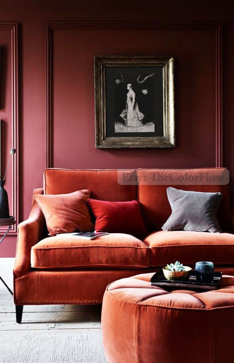 May include: A burnt orange velvet sofa with three cushions, one red, one orange, and one grey. The sofa is in front of a burgundy wall with a framed black and white print. A matching burnt orange velvet footstool sits in front of the sofa.