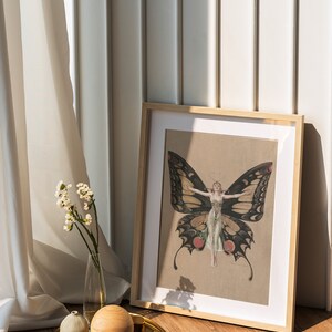 Pode incluir: Uma impress&atilde;o emoldurada de uma mulher com asas de borboleta. A impress&atilde;o est&aacute; em uma moldura marrom clara e est&aacute; sentada em um piso de madeira. H&aacute; dois objetos decorativos em uma bandeja dourada na frente da moldura.