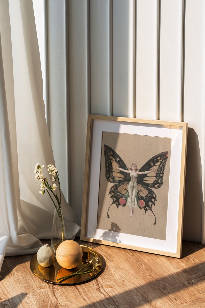Pode incluir: Uma impress&atilde;o emoldurada de uma mulher com asas de borboleta. A impress&atilde;o est&aacute; em uma moldura marrom clara e est&aacute; sentada em um piso de madeira. H&aacute; dois objetos decorativos em uma bandeja dourada na frente da moldura.