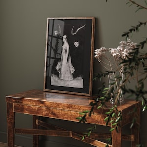 May include: A black and white framed print of a woman in a long dress standing in front of a dark background. The print is on a wooden table with a vase of dried flowers in the foreground.