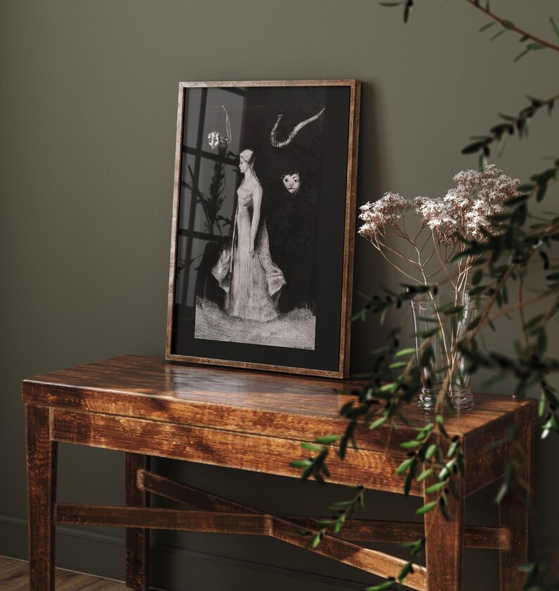 May include: A black and white framed print of a woman in a long dress standing in front of a dark background. The print is on a wooden table with a vase of dried flowers in the foreground.