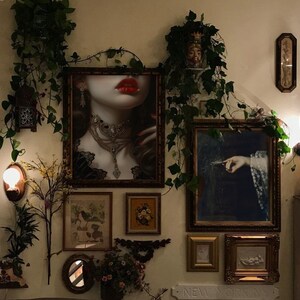Puede incluir: Una galer&iacute;a de pared con una variedad de obras de arte enmarcadas, que incluyen un retrato de una mujer con labios rojos, una pintura de una mano sosteniendo una pluma y una impresi&oacute;n floral. La pared est&aacute; decorada con vegetaci&oacute;n y una peque&ntilde;a l&aacute;mpara.