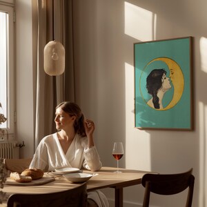 Puede incluir: Una mujer se sienta en una mesa de madera con una copa de vino tinto, un plato de pan y un plato. Un cuadro enmarcado de una mujer besando una luna creciente cuelga en la pared detr&aacute;s de ella.