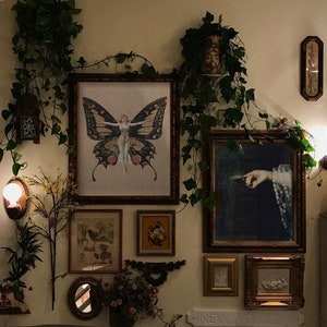 Pode incluir: Uma parede decorada com obras de arte emolduradas, incluindo uma ilustra&ccedil;&atilde;o de borboleta, uma estampa floral e uma pintura de uma m&atilde;o segurando uma pena. A parede tamb&eacute;m &eacute; adornada com vegeta&ccedil;&atilde;o e um pequeno espelho.
