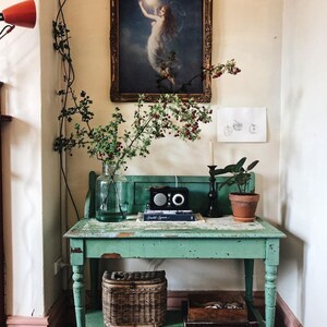 May include: A vintage green table with a distressed finish and a lower shelf. The table has a wicker basket on the shelf and a framed painting of a woman holding a moon above her head. The painting is in a gold frame. There is a small potted plant on the table.