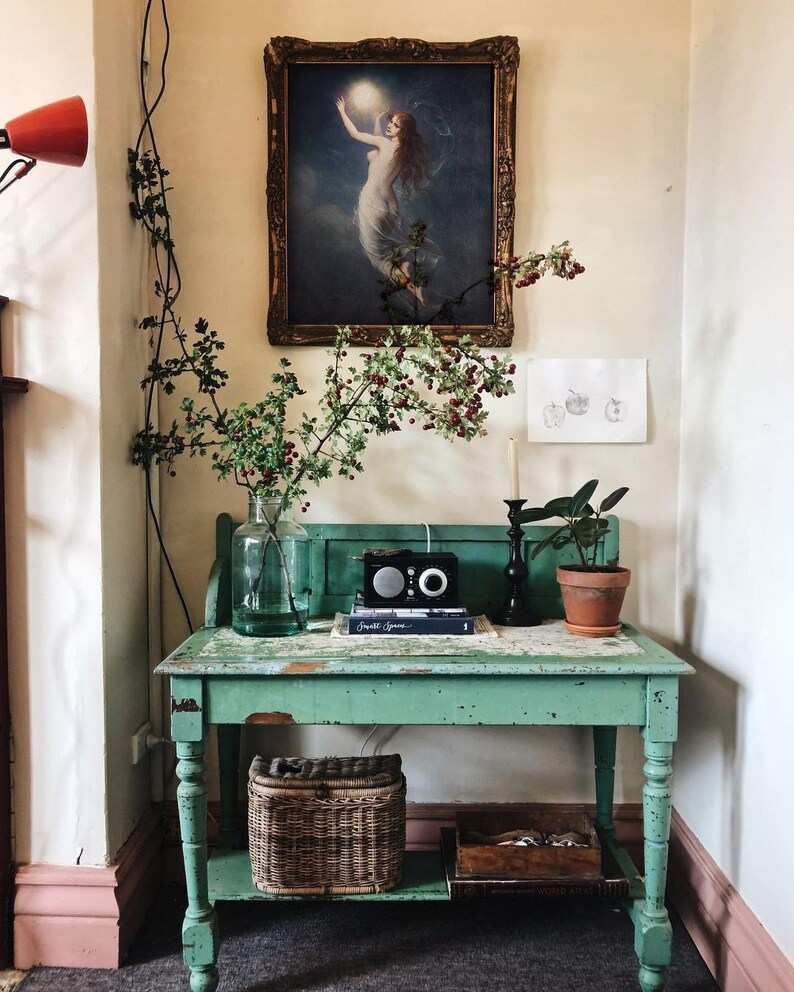 May include: A vintage green table with a distressed finish and a lower shelf. The table has a wicker basket on the shelf and a framed painting of a woman holding a moon above her head. The painting is in a gold frame. There is a small potted plant on the table.