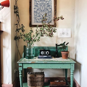 May include: A vintage green table with a lower shelf, featuring a wicker basket, a framed print with a blue and white floral pattern, and a small potted plant. The table has a distressed finish and is decorated with a vase of greenery and a small radio.