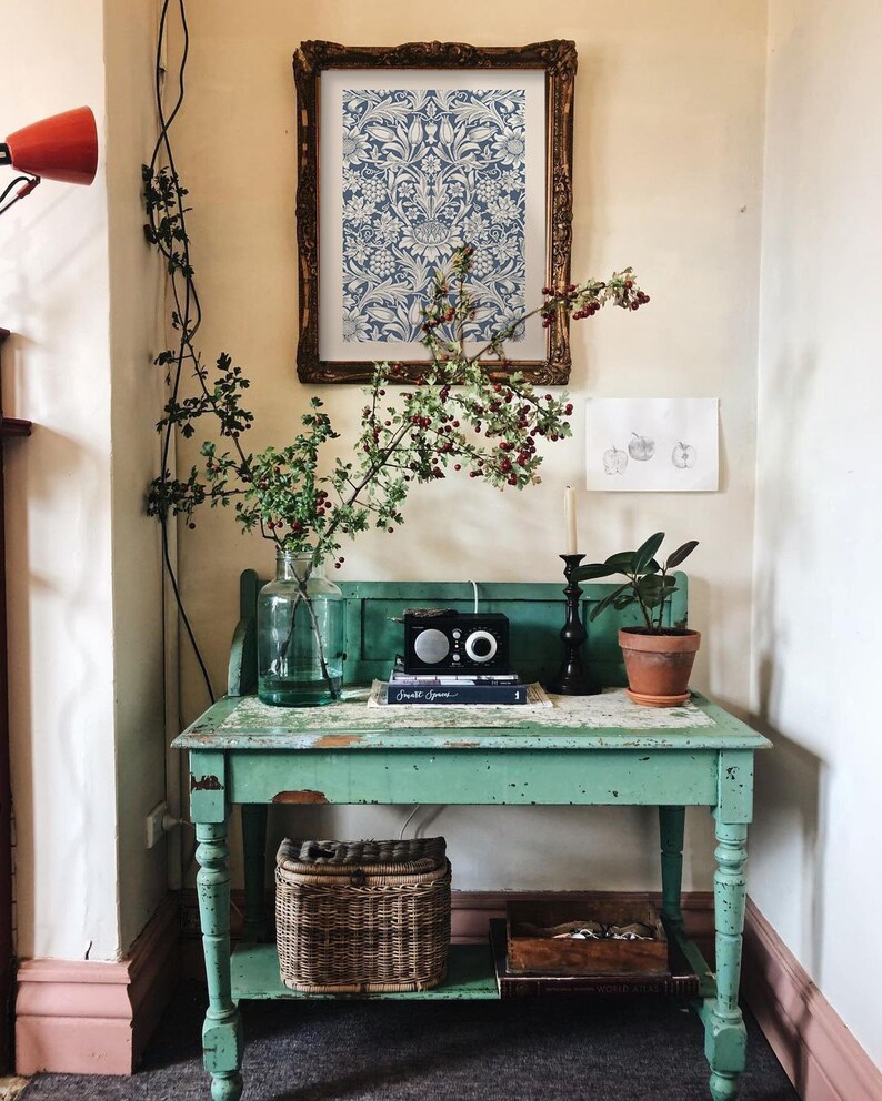 May include: A vintage green table with a lower shelf, featuring a wicker basket, a framed print with a blue and white floral pattern, and a small potted plant. The table has a distressed finish and is decorated with a vase of greenery and a small radio.