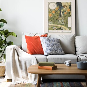 Puede incluir: Un sof&aacute; blanco con una manta, dos almohadas, una naranja y otra azul y blanca con estampado, se encuentra frente a una pared blanca con un p&oacute;ster enmarcado de una mujer en un campo de girasoles. El p&oacute;ster est&aacute; en franc&eacute;s y dice "Aout", que es la palabra francesa para agosto. El sof&aacute; est&aacute; frente a una mesa de centro con dos libros y una taza encima.